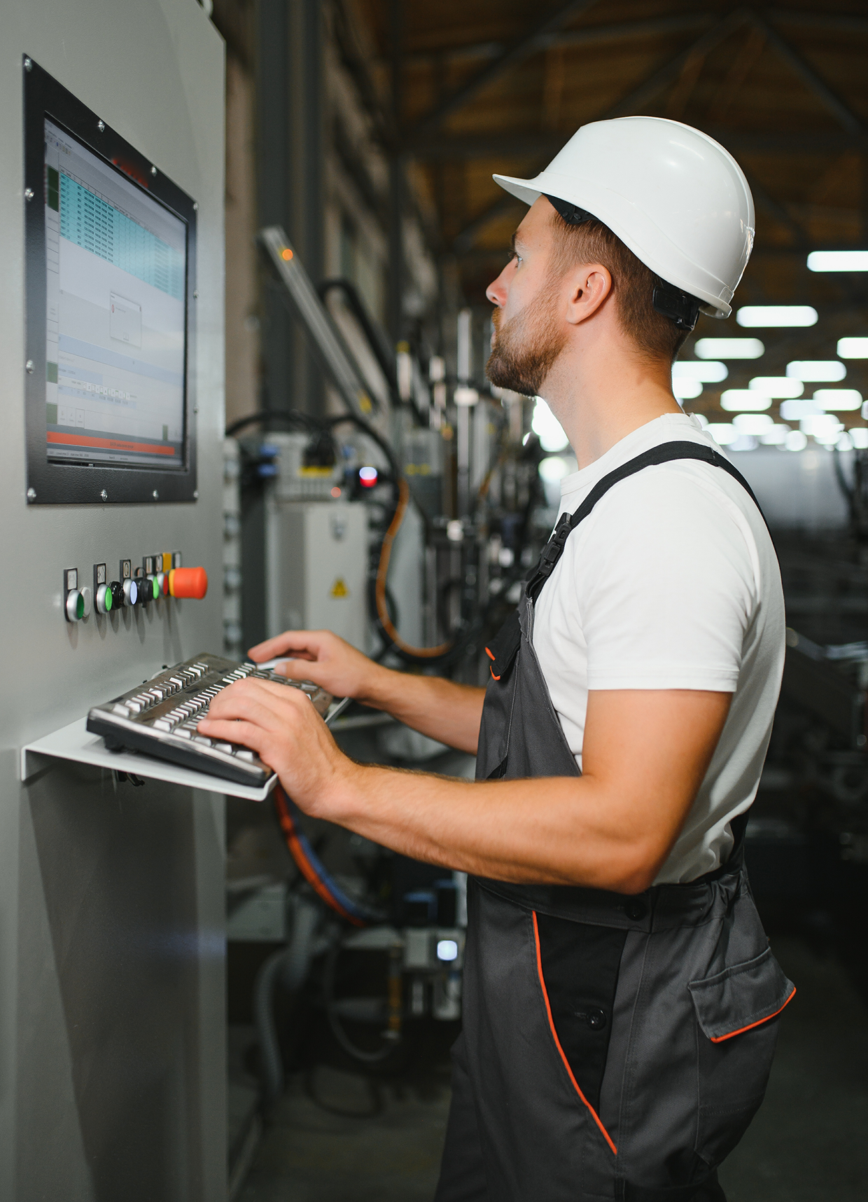A factory worker in a hard hat operating a machine via a computer control panel.